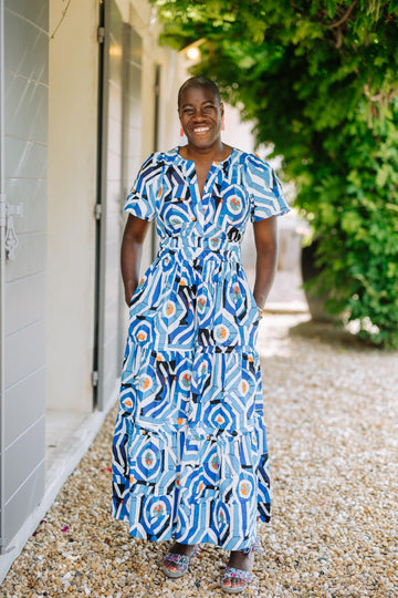 tall dark skinned woman standing outdoors in long dress with blue geometrical patterns under grape leaves next to brick wall